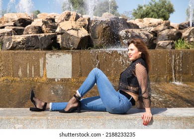 Attractive Mature Woman, Wearing Transparent Black Shirt And Jeans, Sitting Next To A Fountain, Posing In Sensual And Provocative Attitude. Concept Maturity, Beauty, Fashion, Seduction, Provocation.