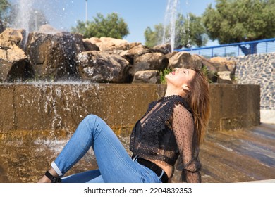 Attractive Mature Woman, Wearing Transparent Black Shirt And Jeans, Sitting Next To A Fountain, Posing In Sensual And Provocative Attitude. Concept Maturity, Beauty, Fashion, Seduction, Provocation.