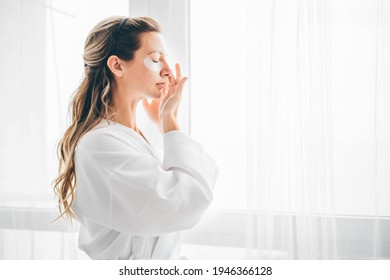 Attractive Mature Woman In White Bathrobe Puts Anti Aging Eye Patch On Face Taking Care Of Skin In Light Modern Hotel Room Close View. 