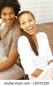 Attractive Merry Young Ethnic Couple Having A Good Laugh While Seated Back To Back On A Brick Patio