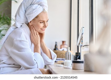 Attractive mid age older adult 50 years old blonde woman wears bathrobe and towel in bathroom touching face, looking at mirror doing daily beauty routine. Skin care treatment concept. - Shutterstock ID 2135514545