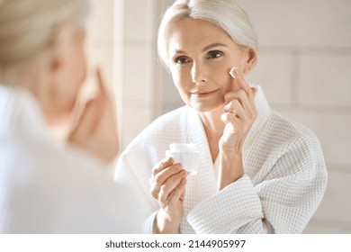 Attractive mid age older adult 50 years old blonde woman wears bathrobe in bathroom applying nourishing antiage face skin care cream treatment, looking at mirror doing daily morning beauty routine.