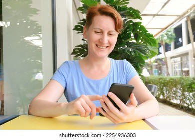 Attractive middle age woman sitting at a table in street cafe, smiles while looking at the phone. Woman looks through information on smartphone and smiles. Communication on the phone, social networks - Shutterstock ID 2254936303