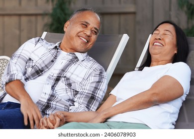 Attractive middle aged Asian couple leans back in lounge chairs and laughs while holding hands - Powered by Shutterstock