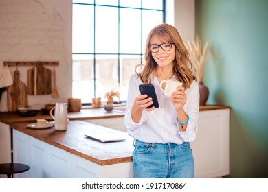 Eine attraktive Frau mittleren Alters, die ihren Morgenkaffee und ihre Textnachrichten trinkt, während sie sich zu Hause in ihrer Küche entspannt. 