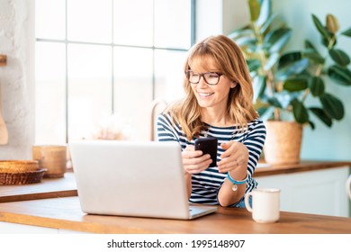 An attractive middle aged woman drinking her morning coffee and text messaging while sitting at home in her kitchen.  - Powered by Shutterstock