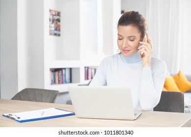Attractive middle aged woman sitting at desk and using her mobile phone and laptop while managing her business from home. Home office. - Shutterstock ID 1011801340