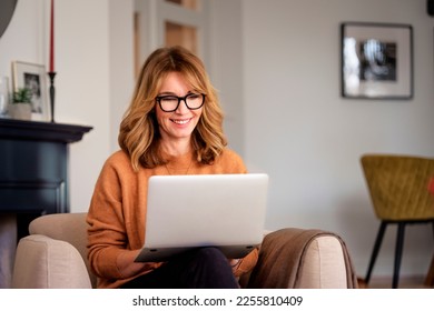 An attractive middle aged woman sitting in the armchair at home and using laptop. Beautiful female wearing casual clothes while working from home. Home office. - Powered by Shutterstock