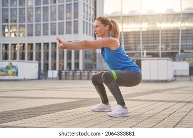 Attractive middle aged woman in sportswear doing squats using resistance band, having workout in the city - Powered by Shutterstock