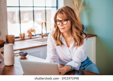 An attractive middle aged woman using her laptop while relaxing at home in her kitchen. 