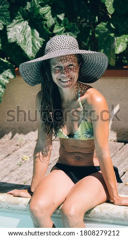Attractive middle aged woman wearing colorful bikini smiling and looking at camera while sunbathing in the swimming pool. She is wearing a sun hat.