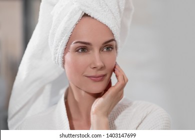 Attractive middle-aged blond woman with white towel on her head and in bathrobe standing in the bathroom by the mirror. She touches skin and smiles. Skin care concept. - Powered by Shutterstock