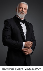 Attractive middle-aged man with grey hair and beard is dressed in a stylish black suit and bowtie, portraying a suave secret agent during a photoshoot against a grey backdrop