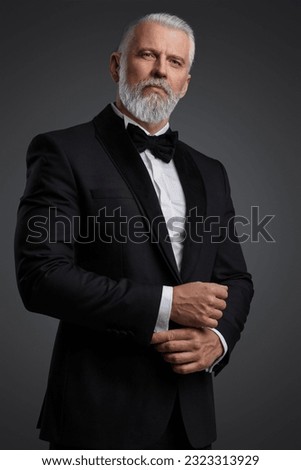 Attractive middle-aged man with grey hair and beard is dressed in a stylish black suit and bowtie, portraying a suave secret agent during a photoshoot against a grey backdrop