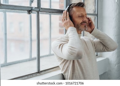 Attractive middle-aged man listening to music on stereo headphones as he leans back against a bright window - Powered by Shutterstock