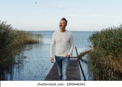 Attraktiver Mann mittleren Alters, der sich auf einem Steg zwischen hohen Schilfrohr auf einem ruhigen See oder Ozean posiert und auf die Seite blickt, während er zur Kamera geht