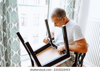 attractive middle-aged man, in white T-shirt, screwing screws in chair with screwdriver, repairing home furniture. Household chores, duties, furniture repair, broken woodwork - Powered by Shutterstock