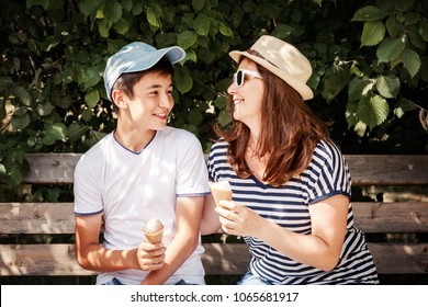 An Attractive Middle-aged Woman With Her Teenage Son Sitting On A Bench And Eating Ice Cream On A Summer Day, Love And A Multinational Family