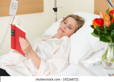 Attractive middle-aged woman lying in a hospital bed propped up on the pillows with a bowl of roses alongside her reading a book - Powered by Shutterstock