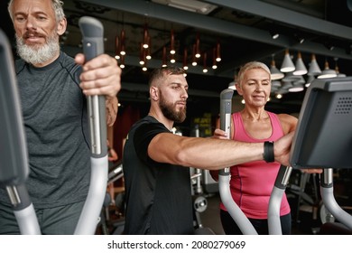 Attractive middle-aged woman with short trendy hairstyle standing on stepper machine waiting young trainer to set up computer. Active woman first time at gym. Mature man excercising near. - Powered by Shutterstock