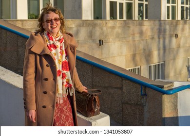 Attractive Middle-aged Woman Wearing Stylish Coat Standing With Bag On Stairs Of Office Building In Early Spring At Sunset. Street City Portrait, Fashion Urban Outfit. Dismissal From Office Concept