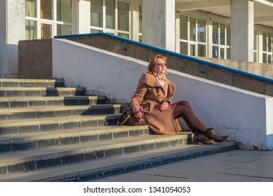 Attractive Middle-aged Woman Wearing Stylish Coat And Shoes Sitting With Bag On Stairs Of Building In Early Spring At Sunset. Street City Portrait, Fashion Urban Outfit. Dismissal From Office Concept