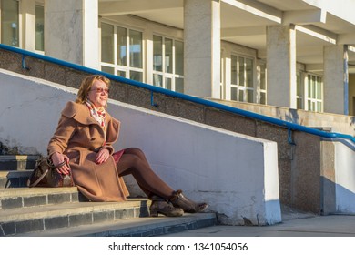 Attractive Middle-aged Woman Wearing Stylish Coat And Shoes Sitting With Bag On Stairs Of Building In Early Spring At Sunset. Street City Portrait, Fashion Urban Outfit. Dismissal From Office Concept