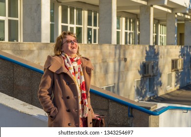 Attractive Middle-aged Woman Wearing Stylish Coat Standing With Bag On Stairs Of Office Building In Early Spring At Sunset. Street City Portrait, Fashion Urban Outfit. Dismissal From Office Concept