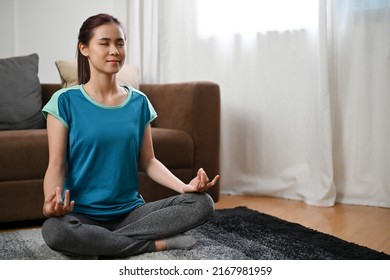 Attractive Millennial Asian Woman Sitting On A Carpet Practice A Yoga At Home, Meditate In Her Living Room.