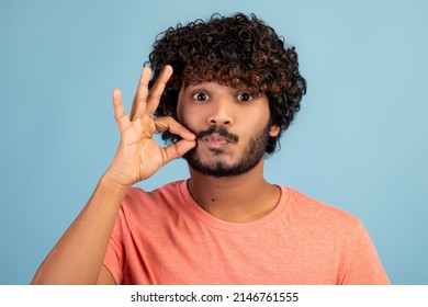 Attractive millennial hindu guy with curly hair keeping secret, showing zipped lips gesture over blue studio background, locking his mouth, closeup photo. Human emotions, gestures concept