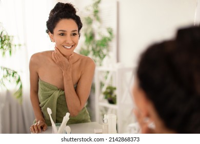 Attractive Millennial Woman In Towel Touching Face, Admiring Radiant Skin Near Mirror At Home, Blank Space. Beautiful Young Female Pampering Herself After Shower At Bathroom