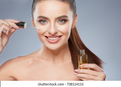 Attractive Model With Brown Hair Fixed Behind, Clean Fresh Skin, Big Eyes And Naked Shoulders Posing At Gray Studio Background, Close Up, Portrait, Holding Face Oil.