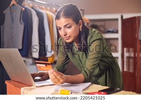Attractive modern young Asian Indian female or woman sitting indoors in front of a laptop typing credit or debit card details or making a digital transaction or payment of festive season shopping.