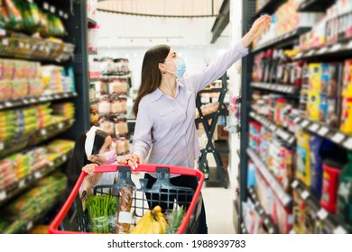 Attractive Mom And Cute Daughter With Face Masks Choosing Products At The Supermarket Aisle And Putting Things In The Shopping Cart 