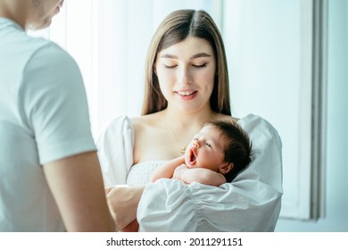 Attractive mother holding cute newborn baby boy in her arms near the window. Cute sleepy baby girl yawning. Happy dad and mom caring for little infant child together.