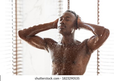 Attractive Muscular Black Man Taking A Shower Washing Body Standing Under Falling Hot Water In Modern Bathroom At Home. Male Bodycare Beauty Routine And Daily Hygiene Concept