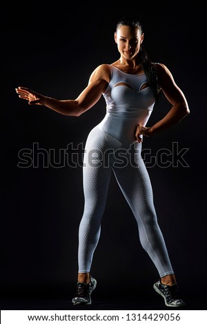 Attractive muscular female in white sportswear keeping hand on waist and looking at camera while standing on tip of toes isolated on black background