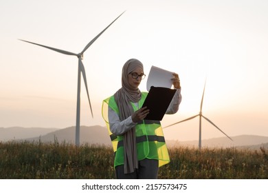 Attractive muslim woman in hijab and eyeglasses looking through some documents on clipboard while standing on farm with windmills. Amazing summer sunset on background.