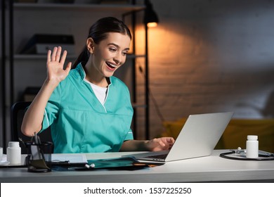 Attractive Nurse In Uniform Sitting At Table And Having Video Chat During Night Shift