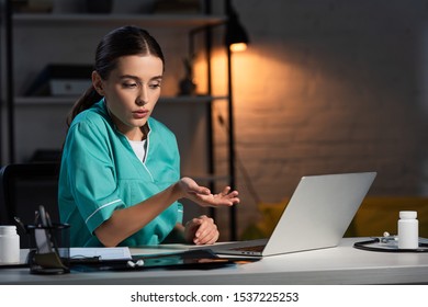 Attractive Nurse In Uniform Sitting At Table And Having Video Chat During Night Shift