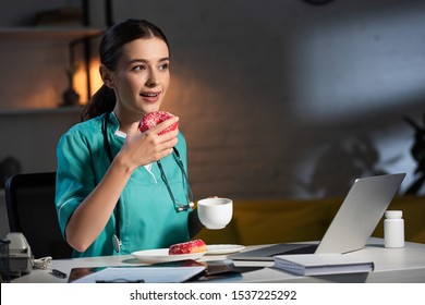 Attractive Nurse In Uniform Sitting At Table And Eating Donut During Night Shift