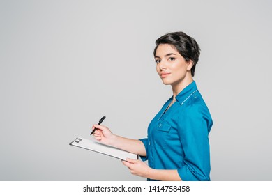 Attractive Nurse In Uniform Writing Ob Clipboard And Looking At Camera Isolated On Grey