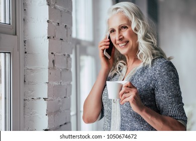 Attractive Old Woman At Home. Senior Smiling Lady Is Sitting On A Sofa With Smart Phone In Hands While Drinking Tea.