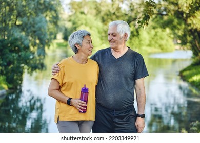 Attractive Older Couple In Sportswear Hugging Pose Look At Each Other Standing In Summer Park Near Lake After Morning Stroll, Sport Active Lifestyle Holds Bottle With Still Water. Healthy Retired Life