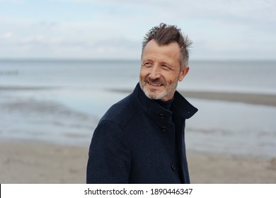 Attraktiver älterer Mann, der an einem kalten Herbsttag entlang eines Sandstrandes geht und mit Interesse und einem ruhigen Lächeln über seine Schulter blickt