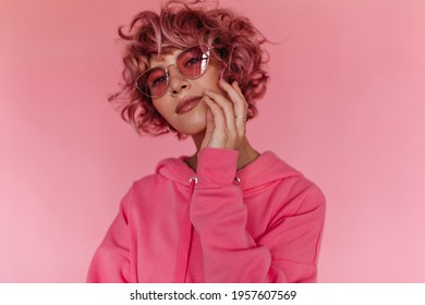 Attractive pink-haired woman in bright colorful sunglasses and fuchsia hoodie looks into camera and touches her face softly on isolated background. - Powered by Shutterstock