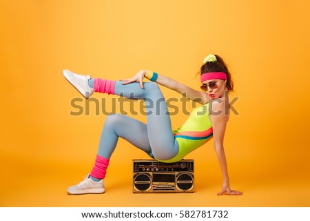 Attractive playful young woman athlete sitting on retro boombox and posing over yellow background
