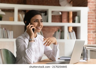 Attractive Pleased Indian Ethnicity Business Lady Sit At Desk With Laptop Holds Smart Phone Lead Business Conversation With Company Client, Enjoy Personal Talk Using Modern Tech, Connection Concept