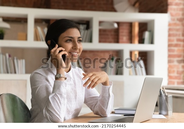 Attractive pleased Indian ethnicity business lady\
sit at desk with laptop holds smart phone lead business\
conversation with company client, enjoy personal talk using modern\
tech, connection\
concept