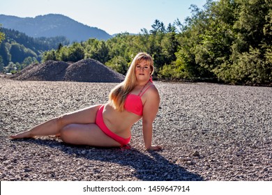 Attractive plus size blond woman in red bikini posing reclining on a pebbly beach in summer sunshine looking at the camera with mountain backdrop